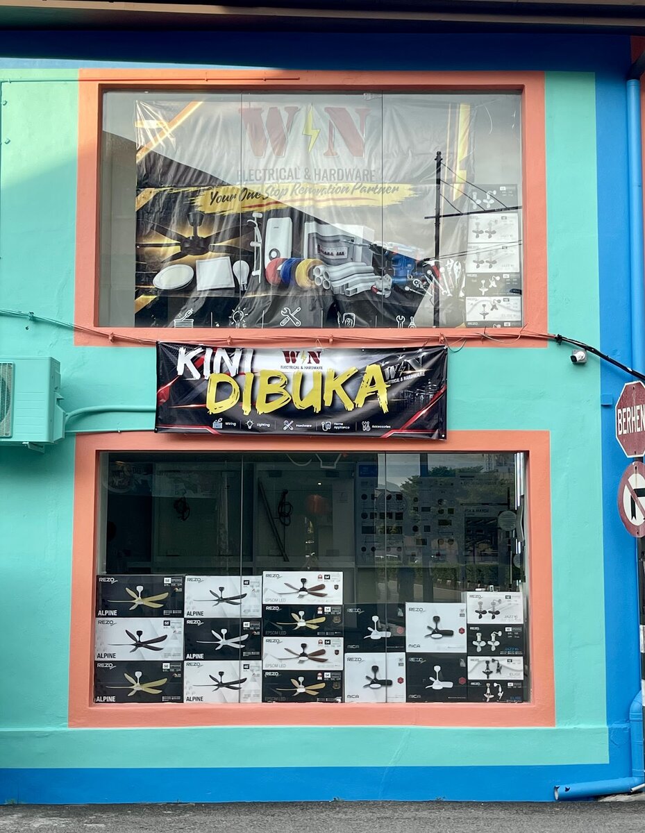 Turquoise and orange shop front with a black Kini Dibuka banner, a Win Electrical & Hardware poster in the upper window and stacked Rezo ceiling fan boxes in the lower window