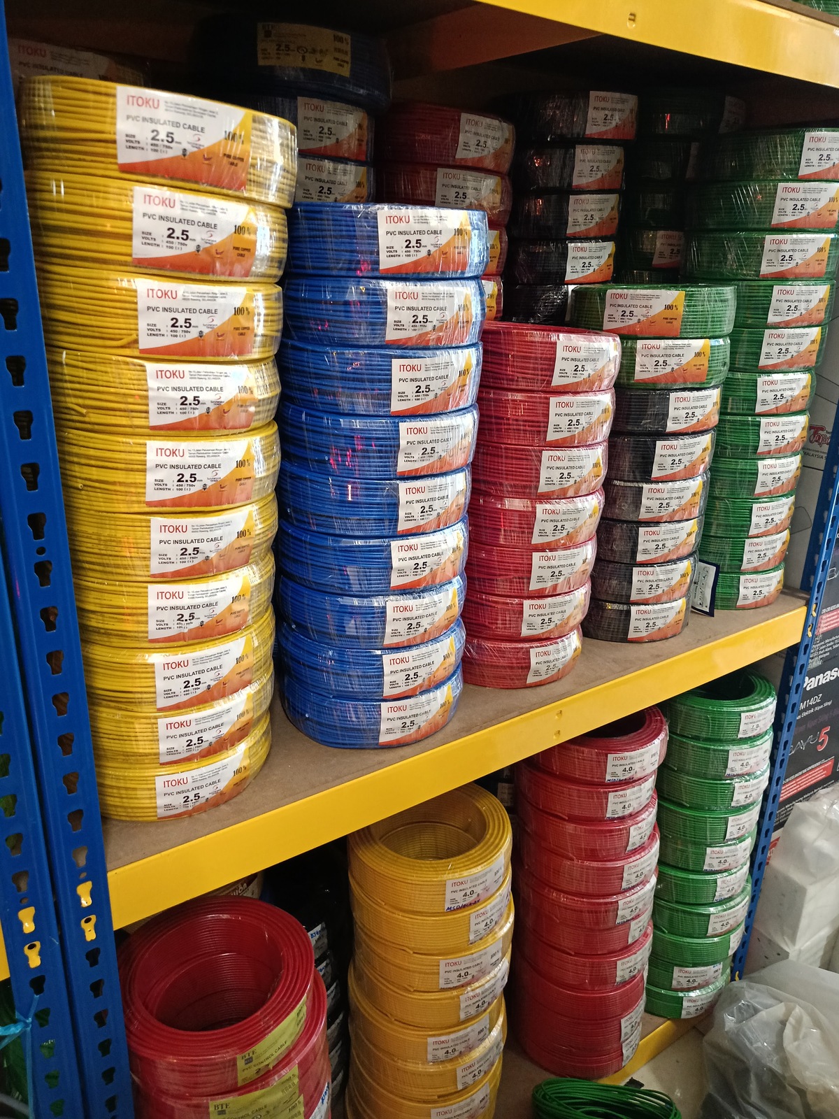 Metal shelving stacked with plastic-wrapped coils of yellow, blue, red, black and green wire, each labelled ITOKU PVC Insulated Cable 2.5