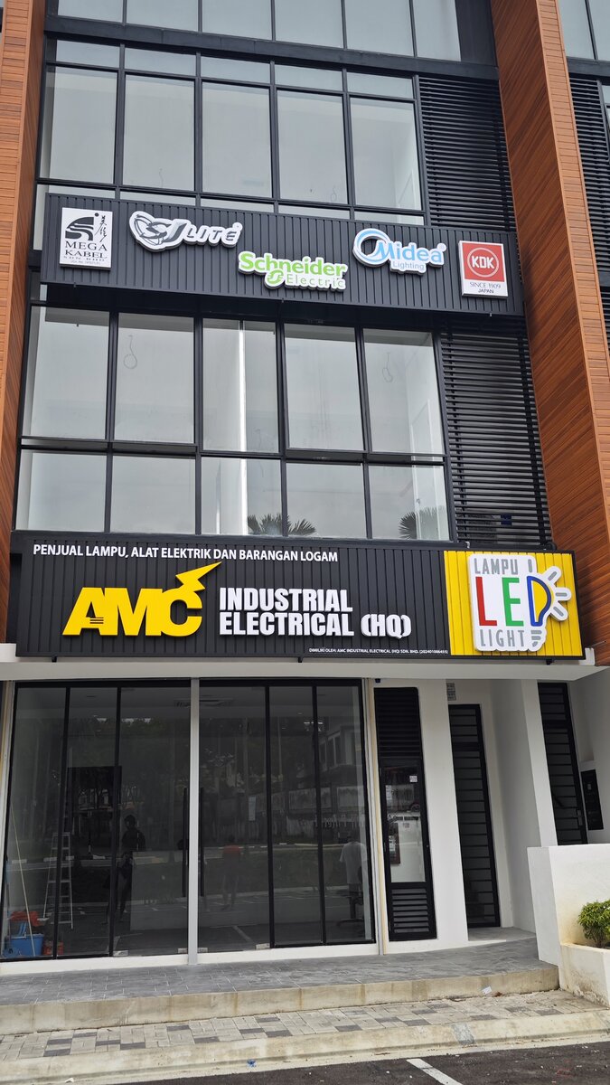 Three-storey shop building with a black AMC Industrial Electrical (HQ) signboard and a yellow Lampu LED Light panel, with brand signs including Schneider Electric and KDK mounted on the upper floor