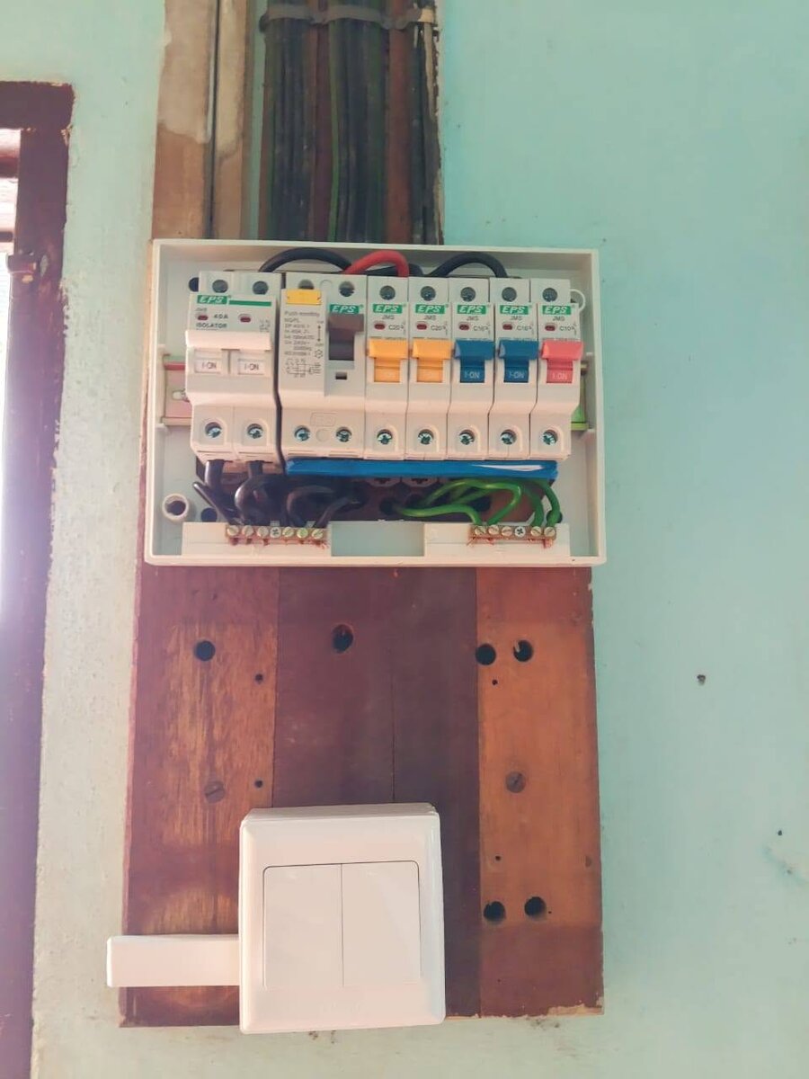 Open white distribution board with EPS isolator, RCCB and five coloured-toggle MCBs, mounted on a dark wooden board with a bundle of cables above and a white two-gang switch below
