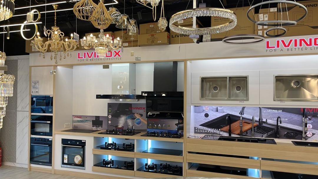 Showroom with crystal chandeliers and pendant lamps hanging above a white kitchen display with ovens, hobs, cooker hoods and stainless-steel sinks under Livinox signs