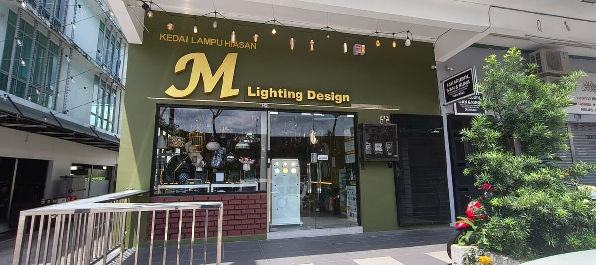 Olive-green shopfront with a gold "M Lighting Design" logo and "Kedai Lampu Hiasan" lettering above glass windows