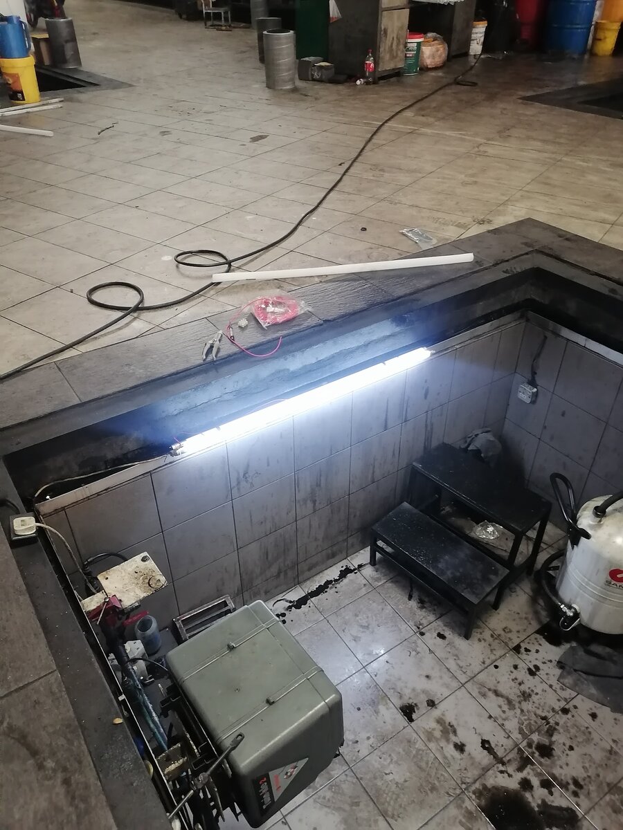 Tiled vehicle service pit in a workshop floor with a lit fluorescent batten along its edge, steps and equipment at the bottom, and a cable running across the floor