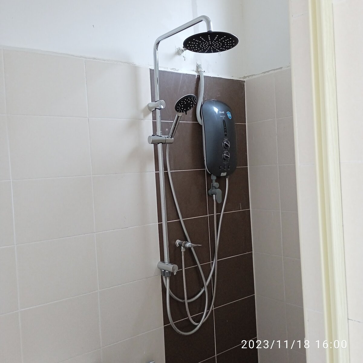 Grey instant water heater mounted on a dark tiled strip in a bathroom, with a rain shower set beside it