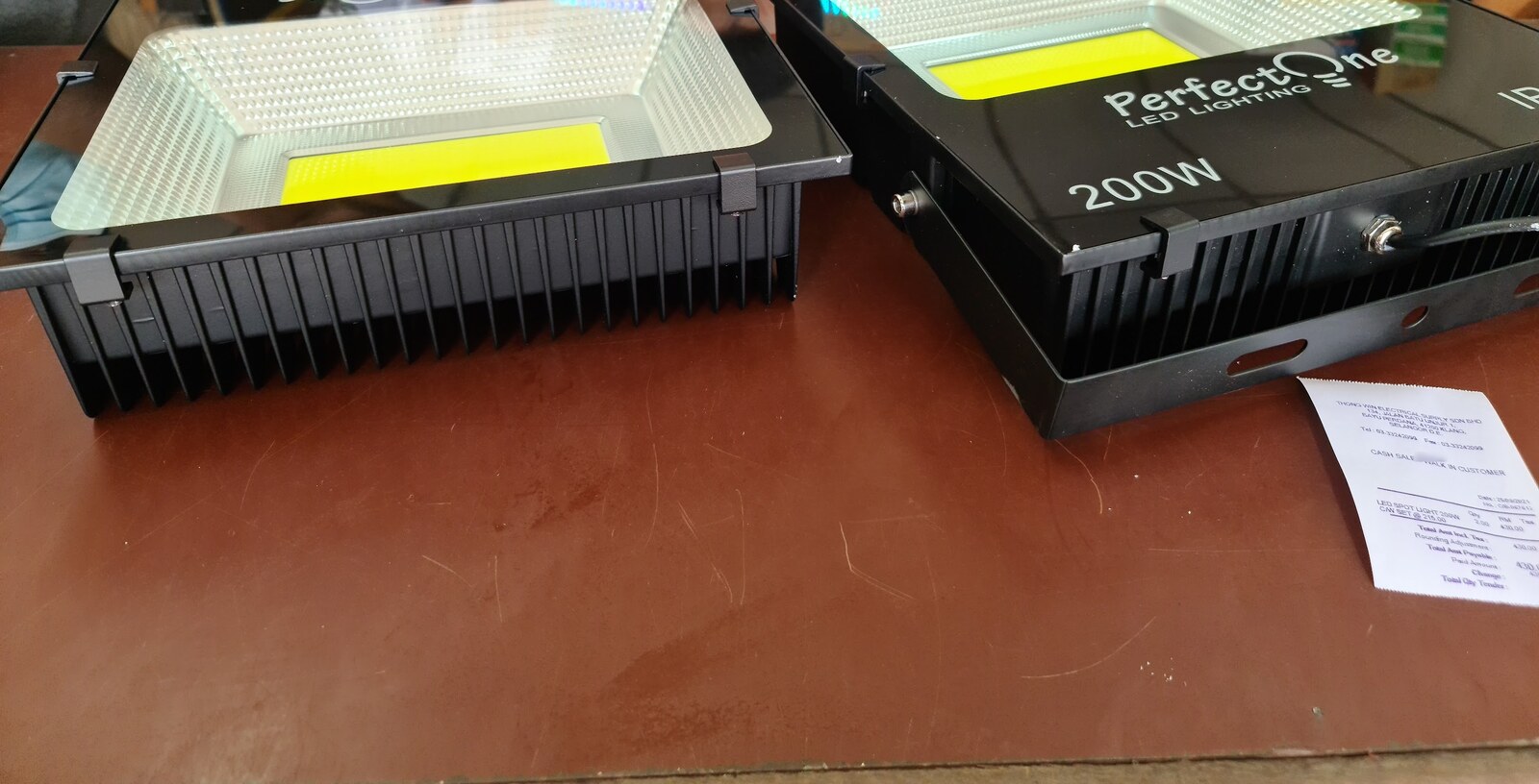 Two black LED floodlights with square yellow LED chips on a brown counter, a printed receipt beside them