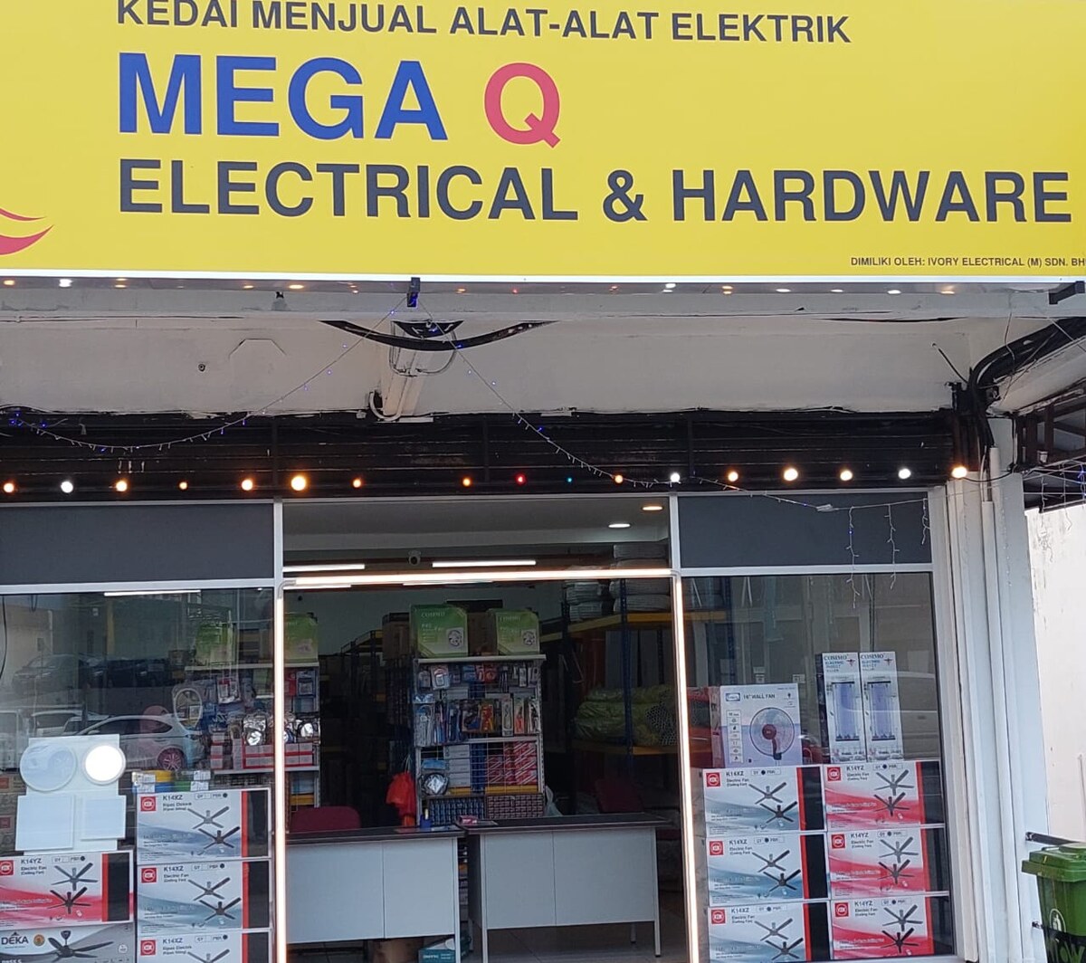 Shopfront under a yellow Mega Q Electrical & Hardware signboard, with stacked KDK ceiling-fan boxes and white insect-killer boxes in the glass window and an open doorway showing a counter and accessory racks