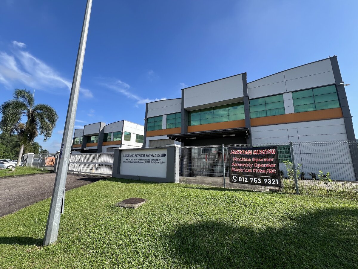 Two-storey grey and orange factory behind a fence, with a white company signboard and a black recruitment banner on the fence