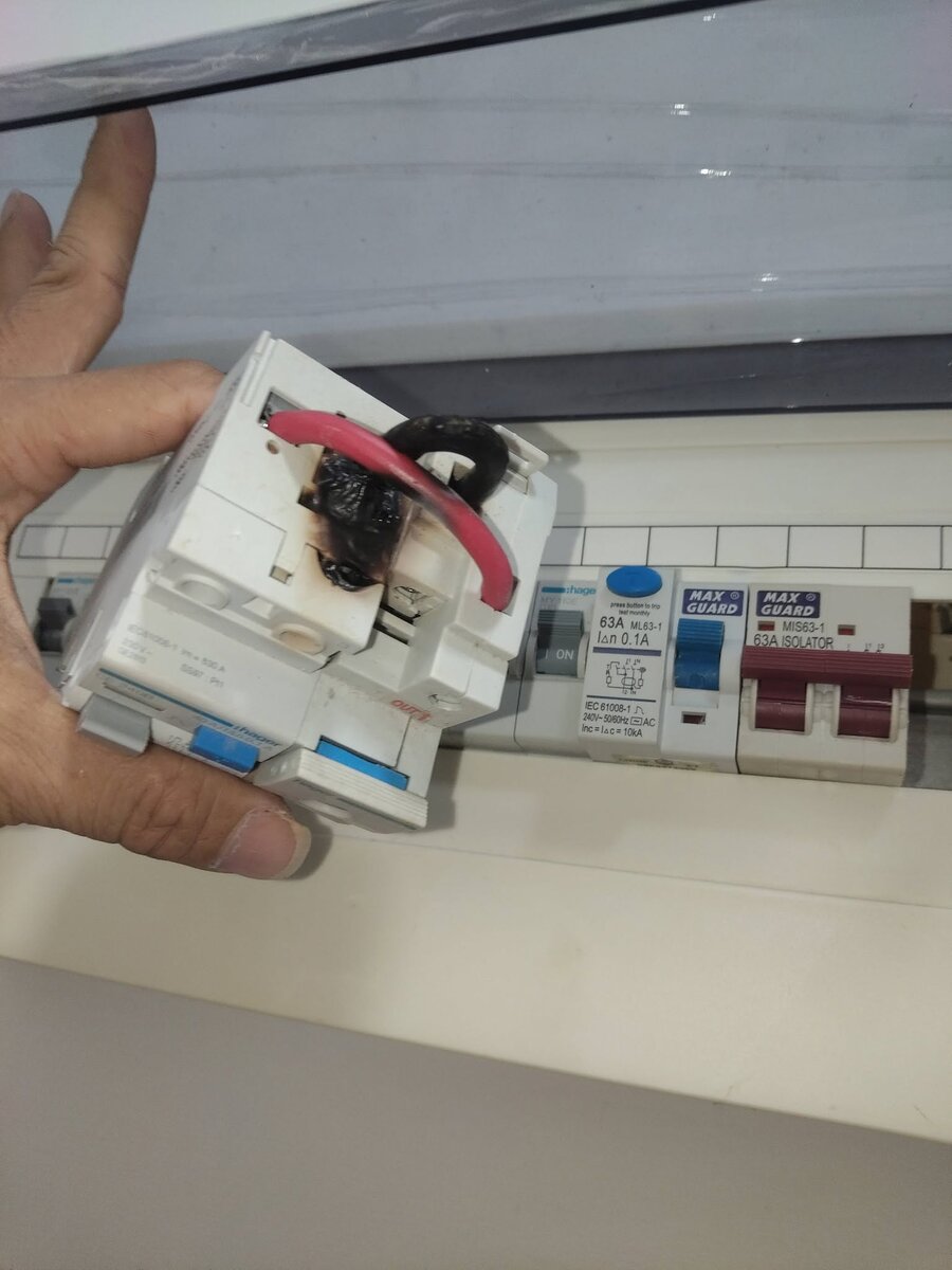 Hand holding a white Hager RCCB with a melted, blackened terminal and a red cable, in front of an open distribution board with a Hager device, a blue-toggle Maxguard RCCB and a Maxguard isolator