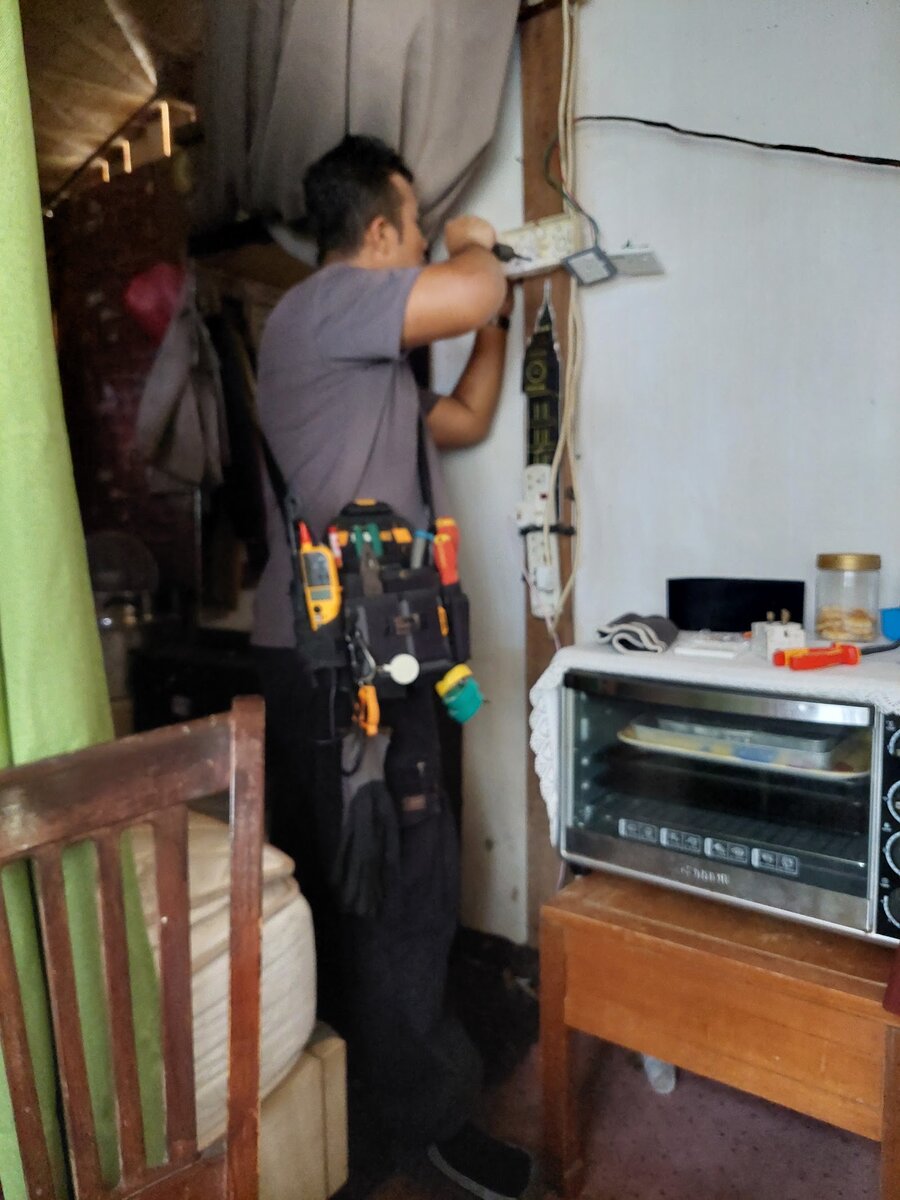 A man in a grey T-shirt with a tool belt working on a wall-mounted socket beside a curtain, with a toaster oven on a small table next to him