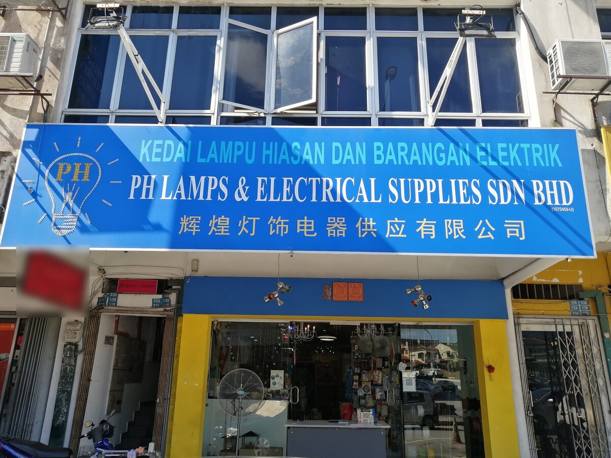 Blue signboard with a light-bulb logo reading PH Lamps & Electrical Supplies Sdn Bhd and Chinese characters over a glass shopfront with chandeliers, a stand fan and a rack of carded accessories inside
