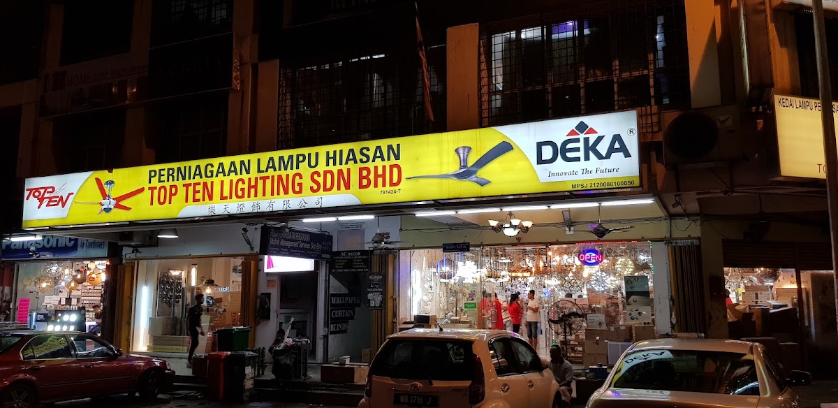 Night view of a yellow Top Ten Lighting signboard with a Deka fan logo above a brightly lit shop full of chandeliers