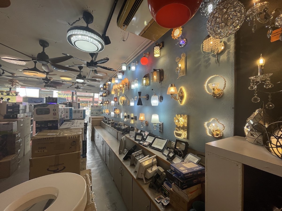 Long narrow showroom with dozens of lit wall lamps on a blue wall, ceiling fans and round ceiling lights above, floodlights on a counter and stacked cartons on the left
