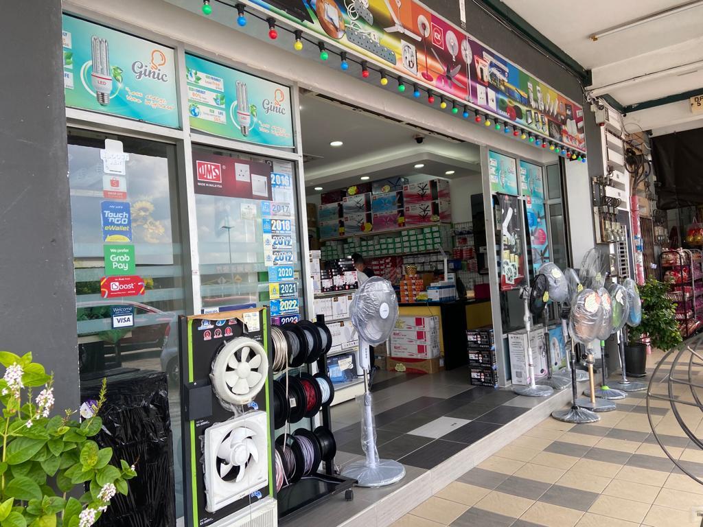 Glass shopfront under a five-foot way with stand fans, exhaust-fan samples and a rack of cable reels outside, and shelves of boxed lights inside