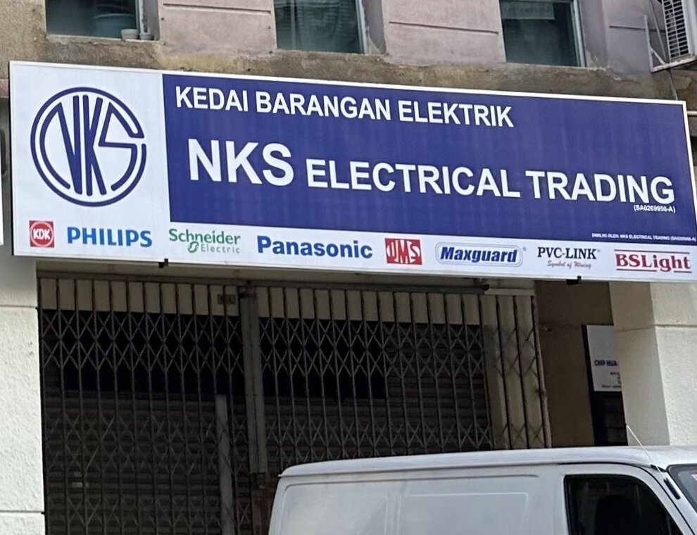 Blue and white shop signboard reading NKS Electrical Trading with a row of brand logos underneath, above a closed grille shutter