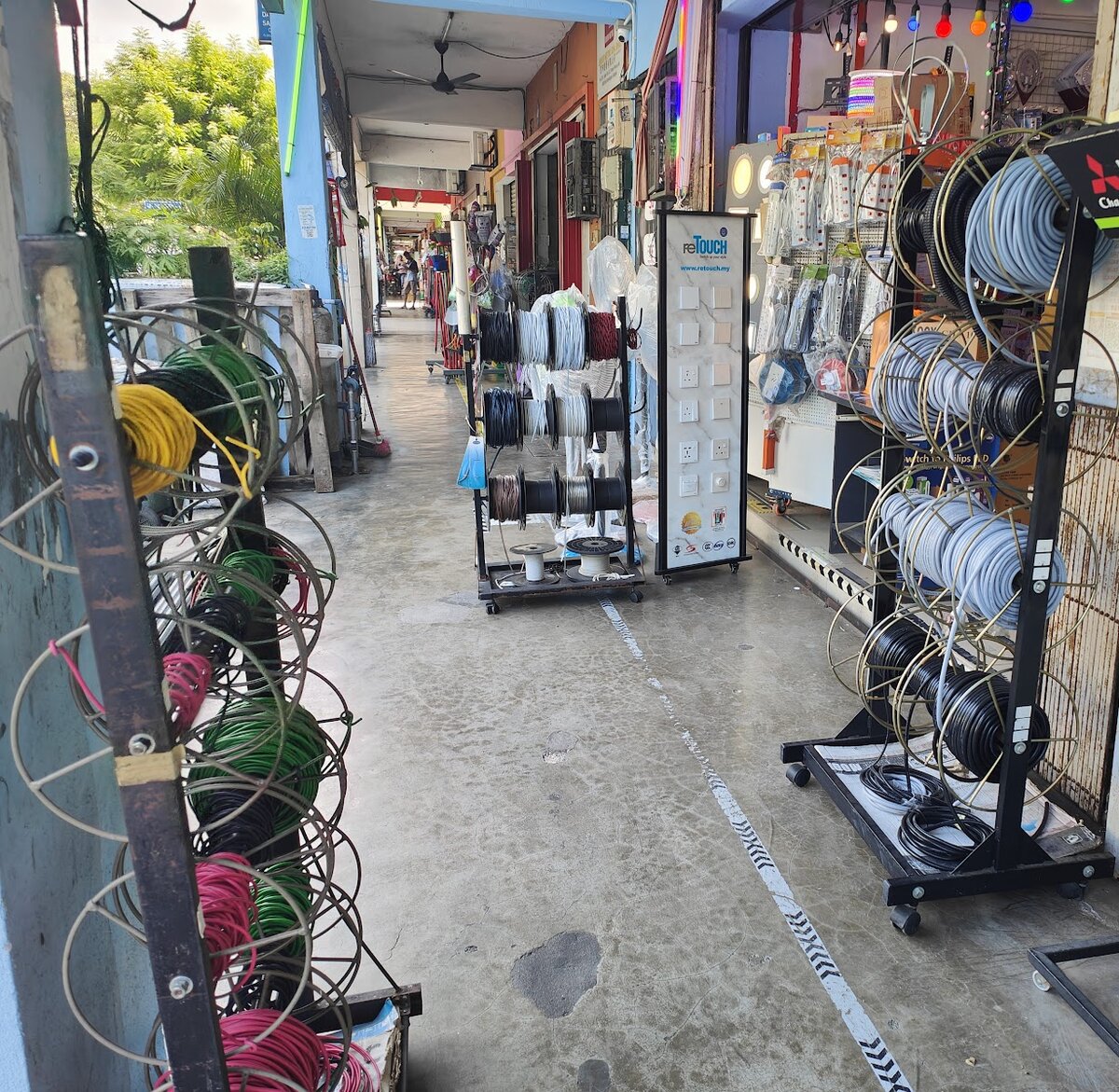 Covered walkway outside a shop lined with metal racks holding coils of coloured and grey cable, a rack of cable reels and a tall white switch display stand