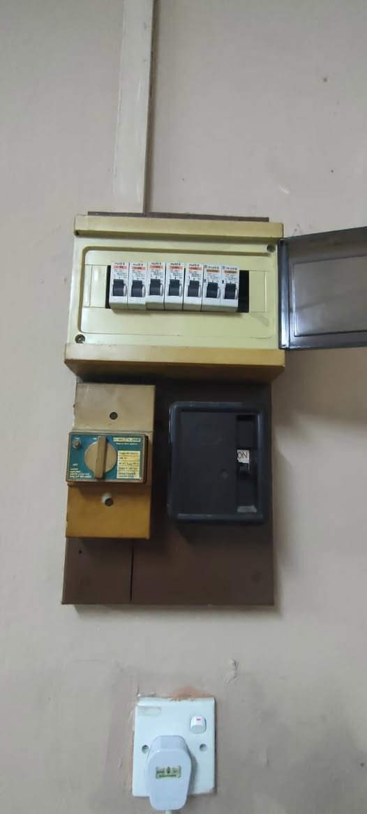 Small cream distribution board with seven breakers, above a wooden board holding an older rotary earth-leakage breaker and a black-covered switch, with a wall socket below