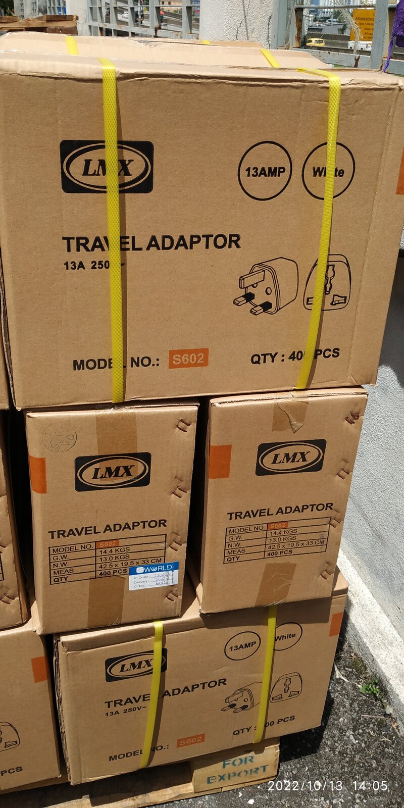 Stack of brown cartons printed with the LMX logo, Travel Adaptor 13A and model number S602, bound with yellow straps on a wooden pallet