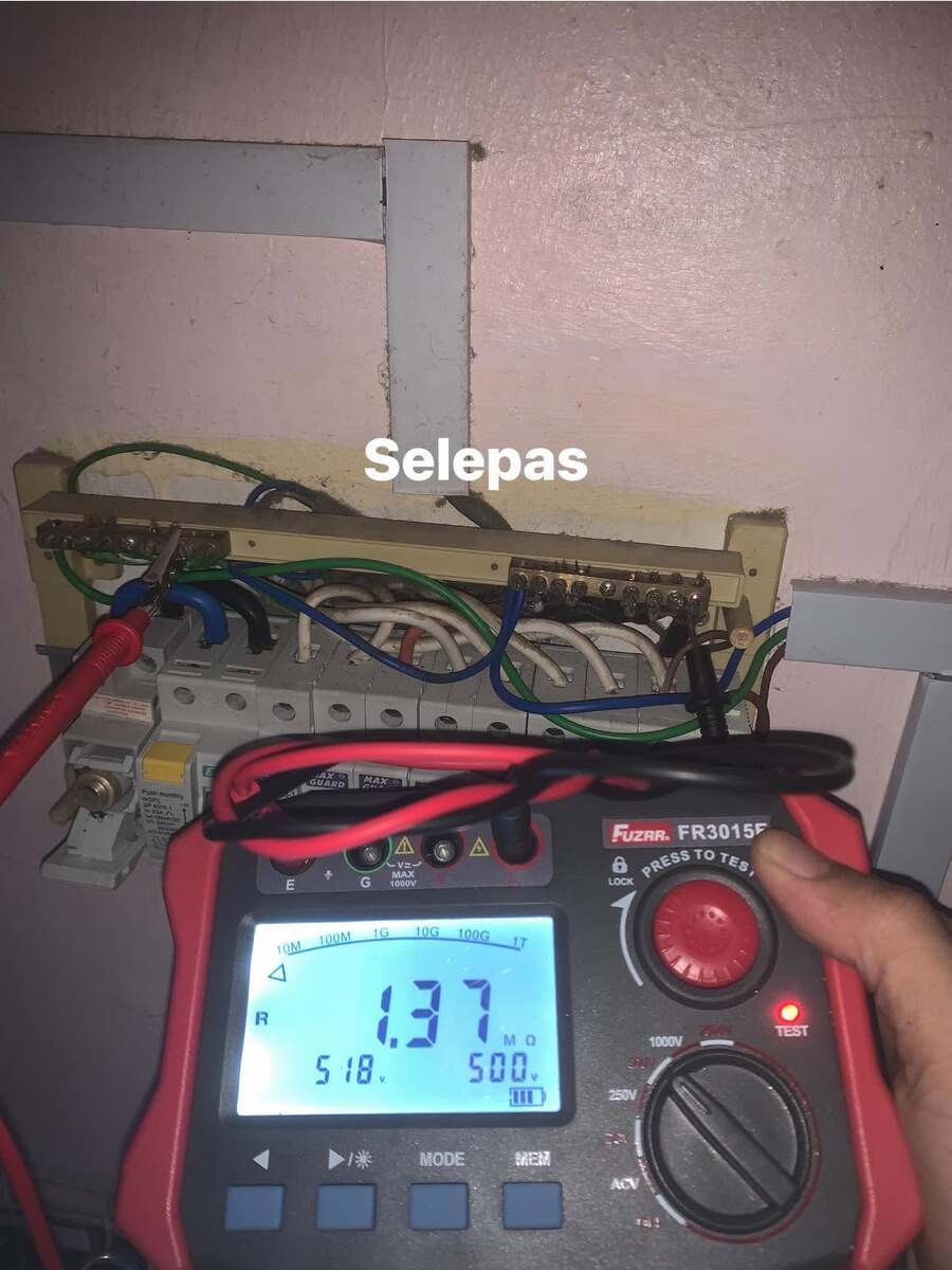 Hand holding a red Fuzrr FR3015E insulation tester whose screen reads 1.37 MΩ and 500 V, test leads clipped to a small DB with a cream backboard on a pink wall with MCBs and coloured wiring, with the word Selepas overlaid