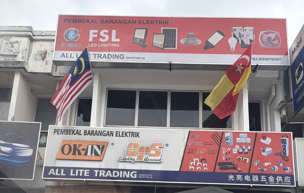 Two-storey shophouse with a red upper signboard advertising Iwachi and FSL LED lighting and a lower signboard reading OK-IN, OPS and All Lite Trading with pictures of electrical parts
