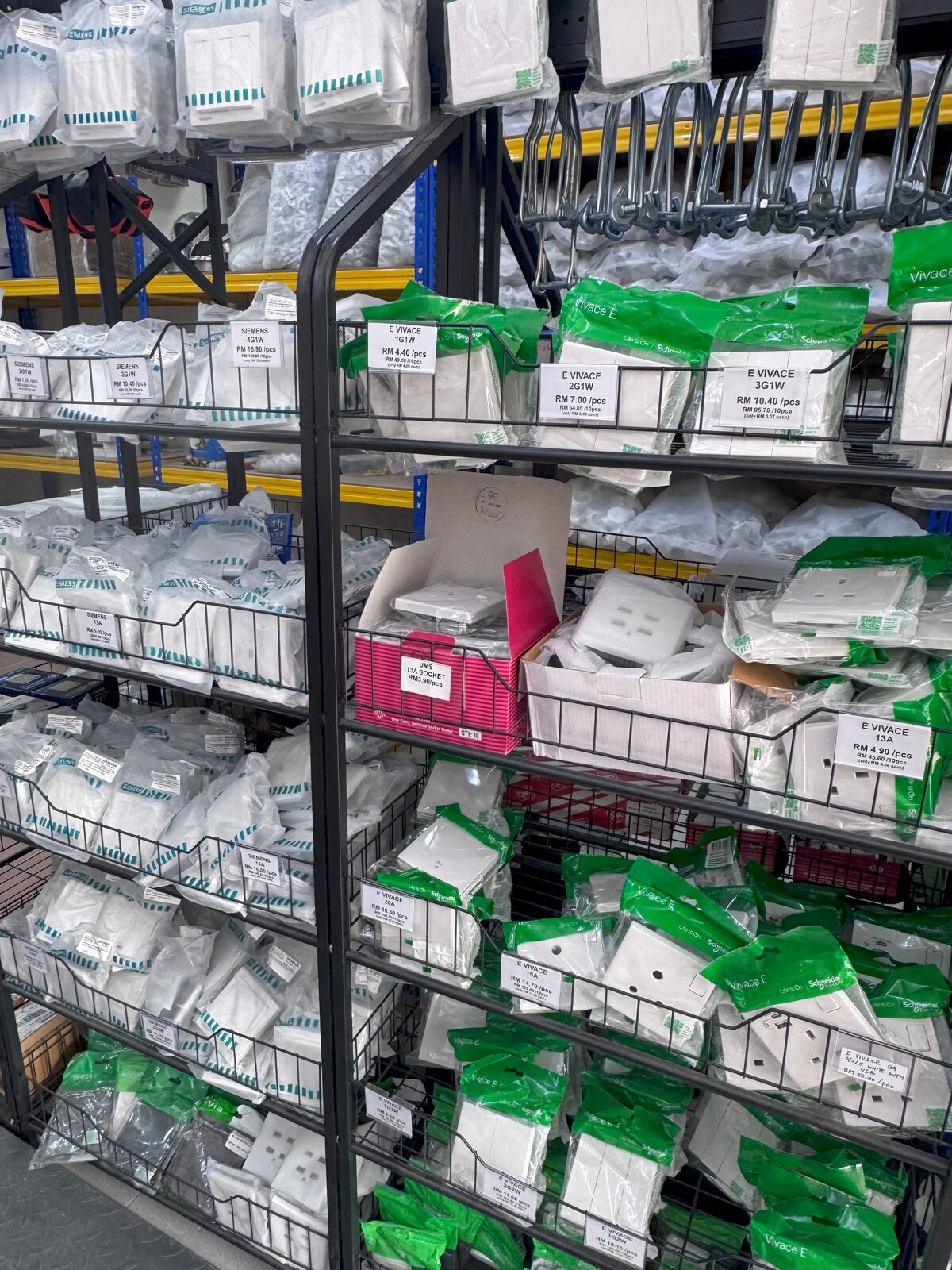 Black wire-basket racks full of white switch and socket packs in Siemens and green Schneider Vivace E bags, each basket with a printed price label