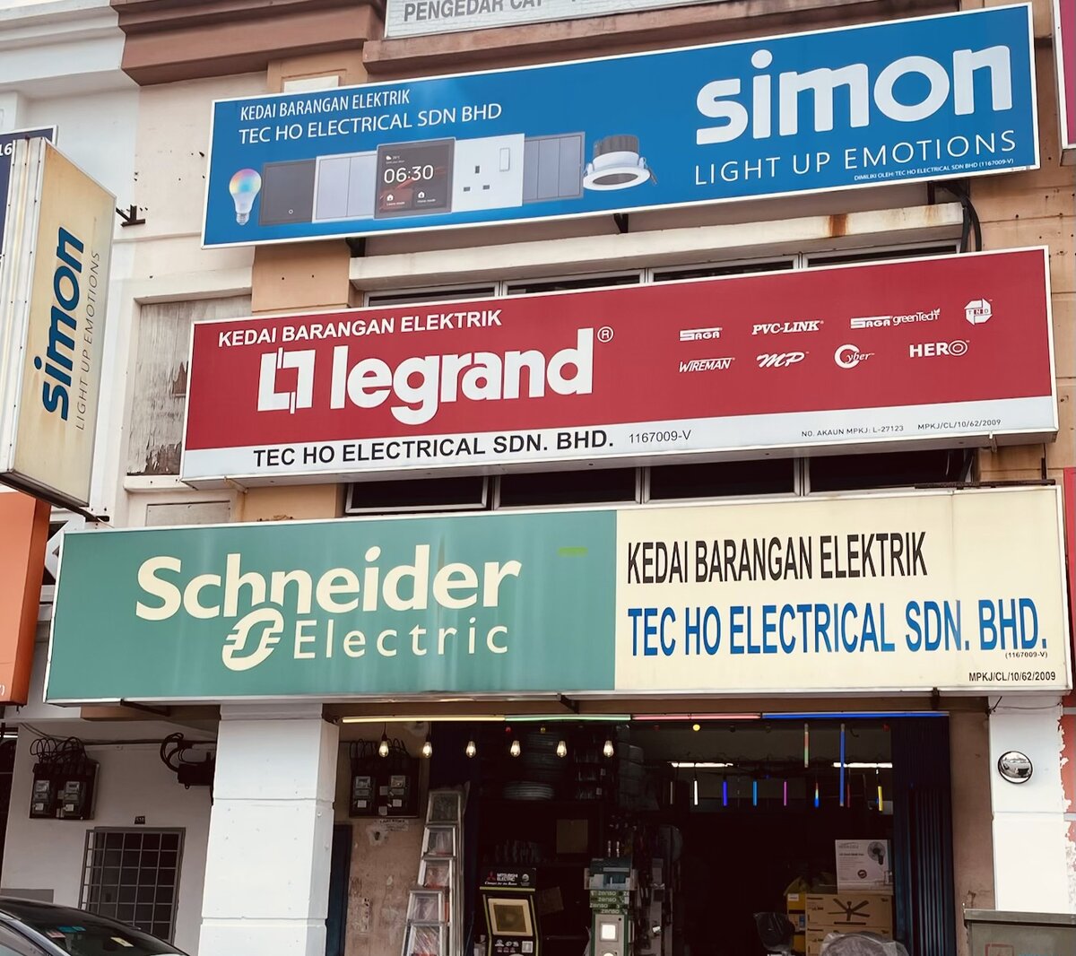 Three stacked shop signboards, blue Simon, red Legrand with small brand logos, and green Schneider Electric with Tec Ho Electrical Sdn. Bhd., above an open shopfront with ladders and fans